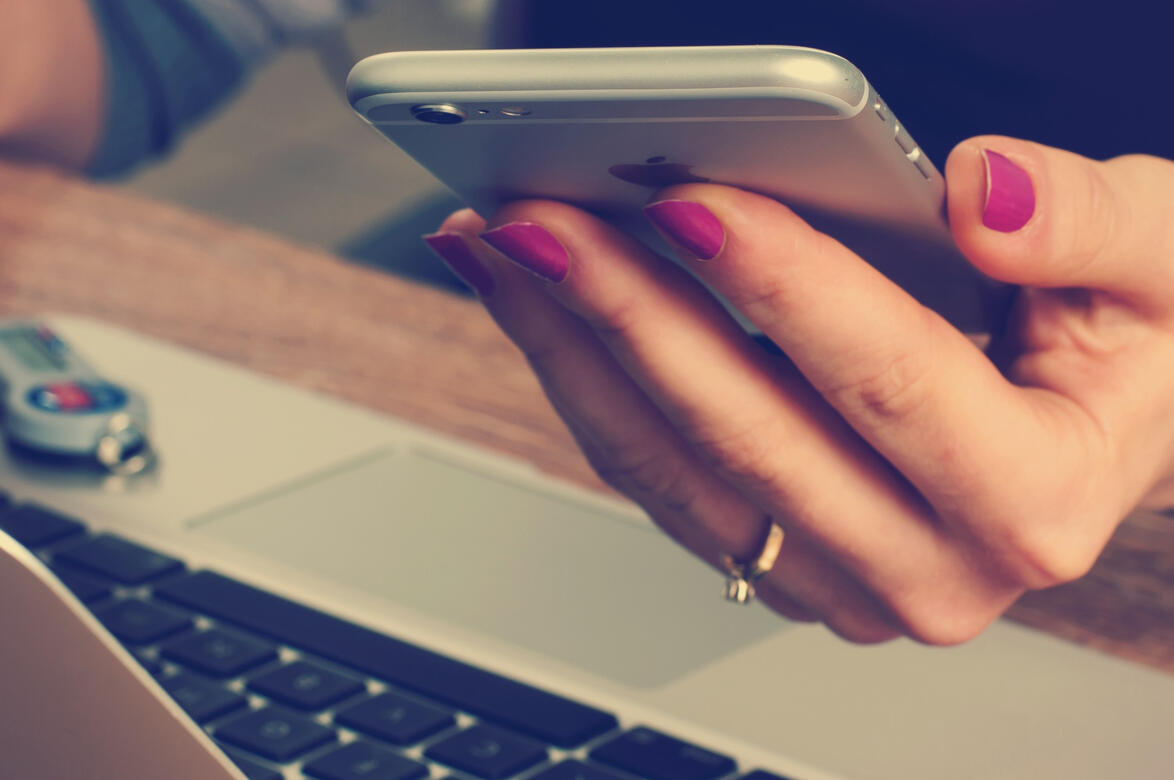 A woman with pink fingernails holding a cell phone in her left hand while sitting in front of a laptop.